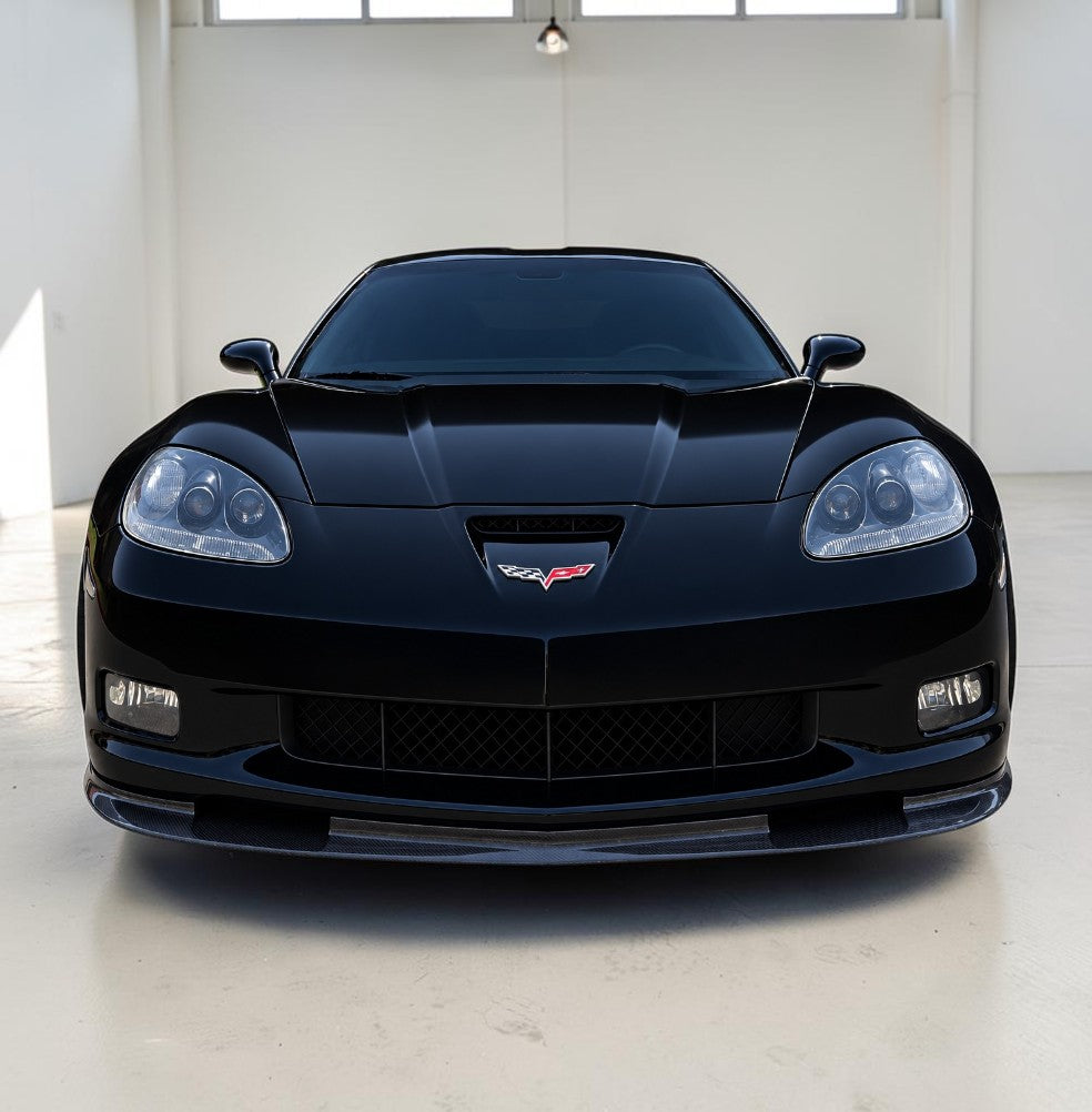 A black Corvette sports car photographed head-on in a clean indoor studio or showroom with white walls and floor.