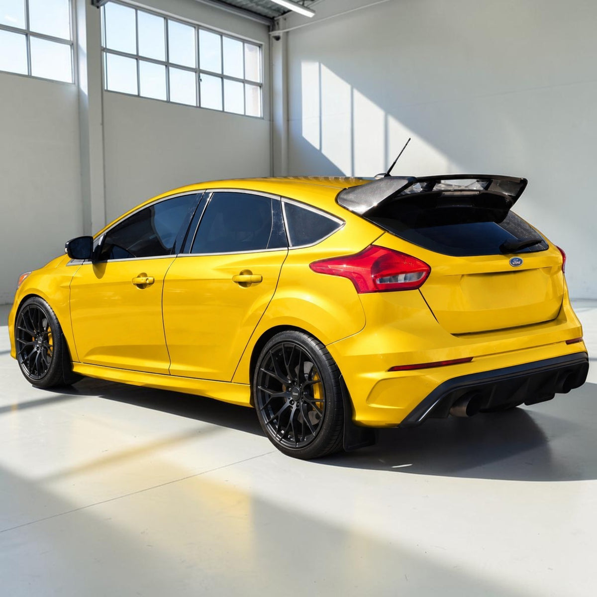 A bright yellow Ford Focus RS hatchback with black wheels, a black rear spoiler, and a black trunk lid, photographed in a clean, sunlit indoor garage or studio setting.
