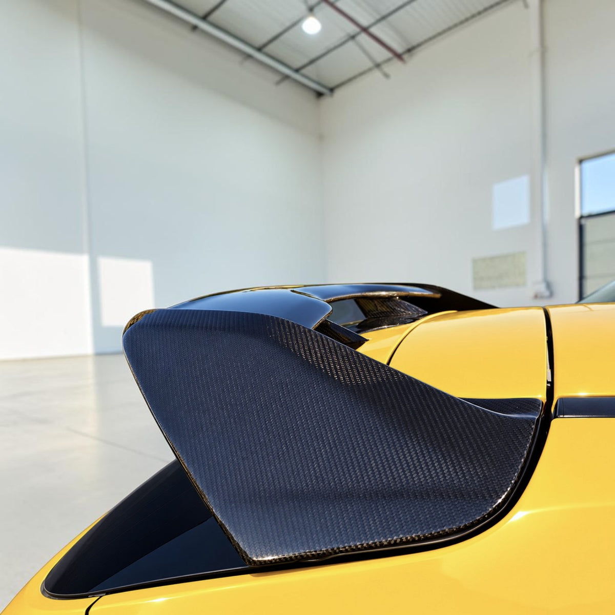 Close-up of a yellow sports car rear section showing a large black carbon-fiber rear spoiler in a bright indoor showroom or garage.