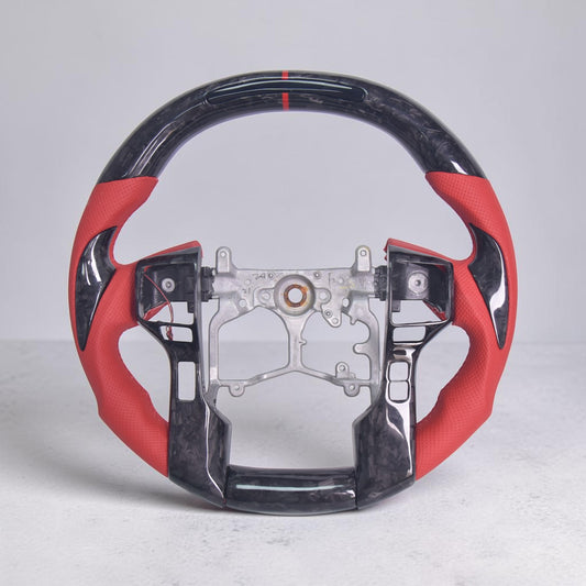 A carbon fiber racing steering wheel with red leather-like grips and a silver metallic center hub, photographed against a neutral light gray background.