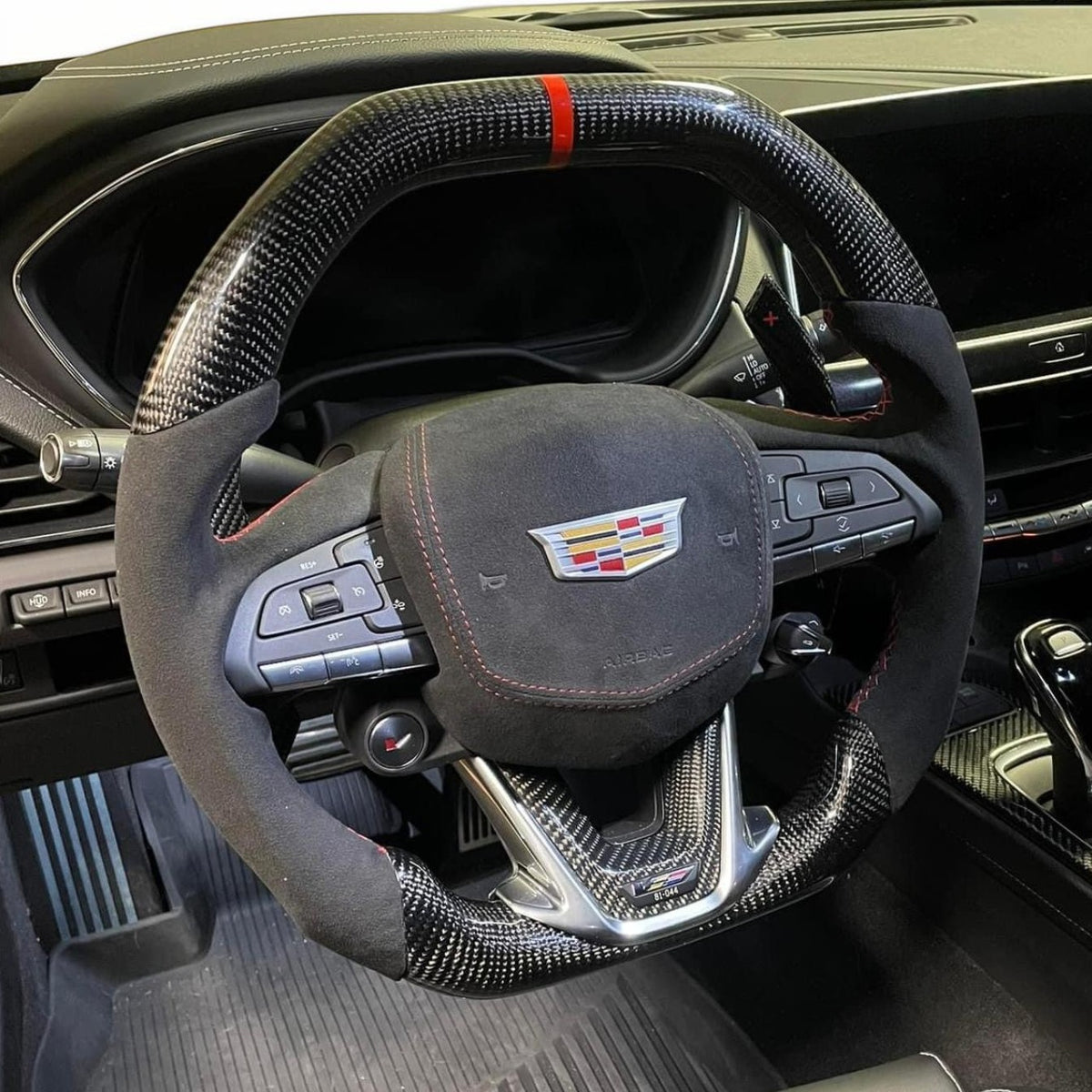 A carbon fiber and suede steering wheel cover installed on a Cadillac steering wheel inside a car cabin. The cover features a black carbon fiber weave pattern, black suede sections, and red stitching accents, including a red stripe on the top rim.