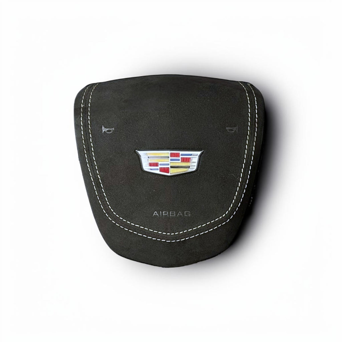 A black suede or nubuck airbag cover featuring white contrast stitching, a metallic Cadillac crest badge, and the word 'AIRBAG' printed below, shown on a white background with a soft drop shadow.