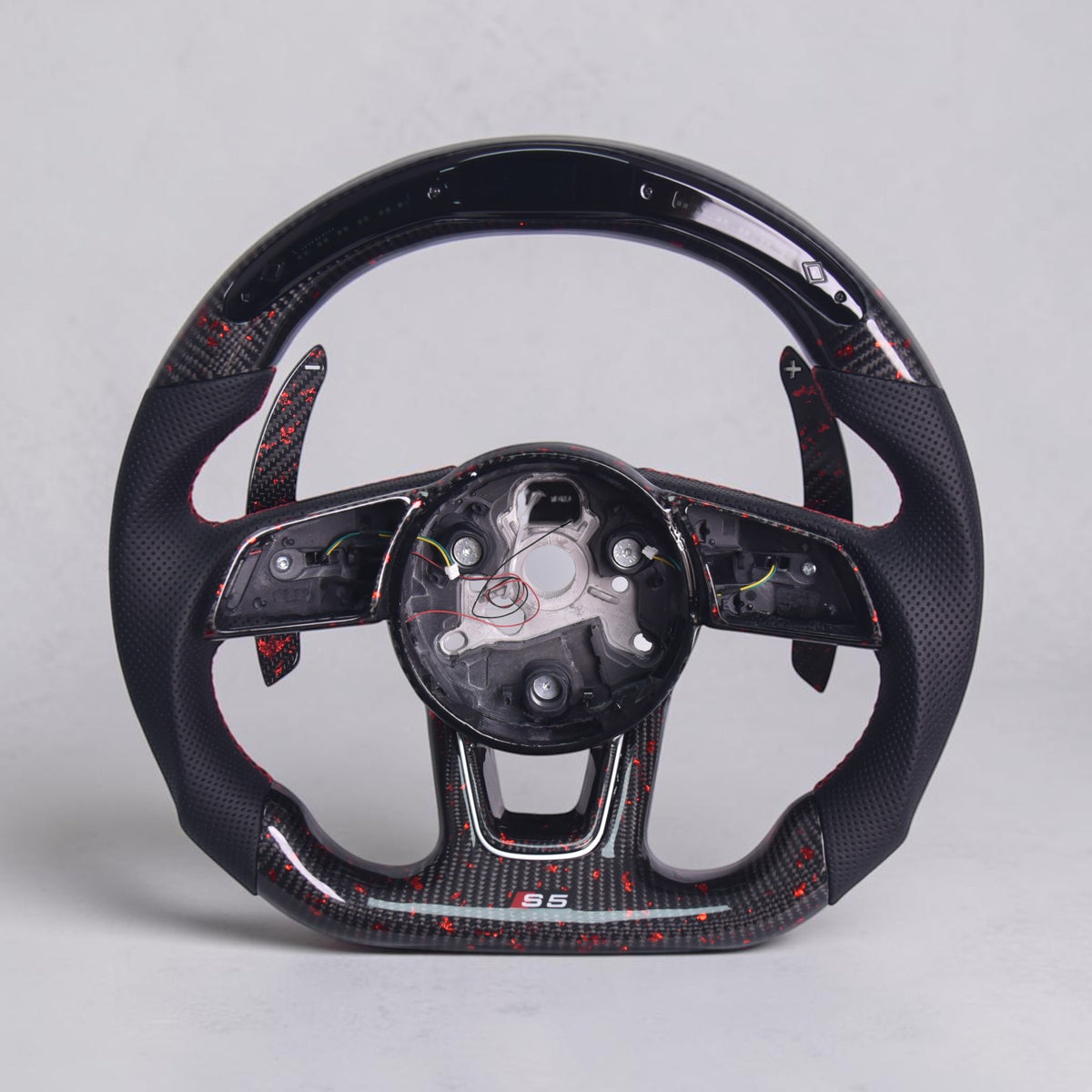 A carbon fiber sports steering wheel with a glossy black finish, red stitching, and red carbon fiber weave patterns, shown empty with the center console removed against a light gray background.